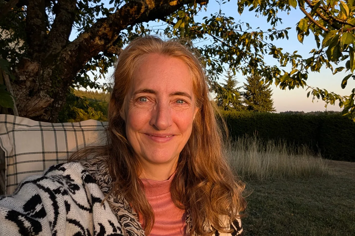 Marianne im Abendlicht unter dem Kirschbaum in ihrem Garten, dahinter Wiese und Wald.